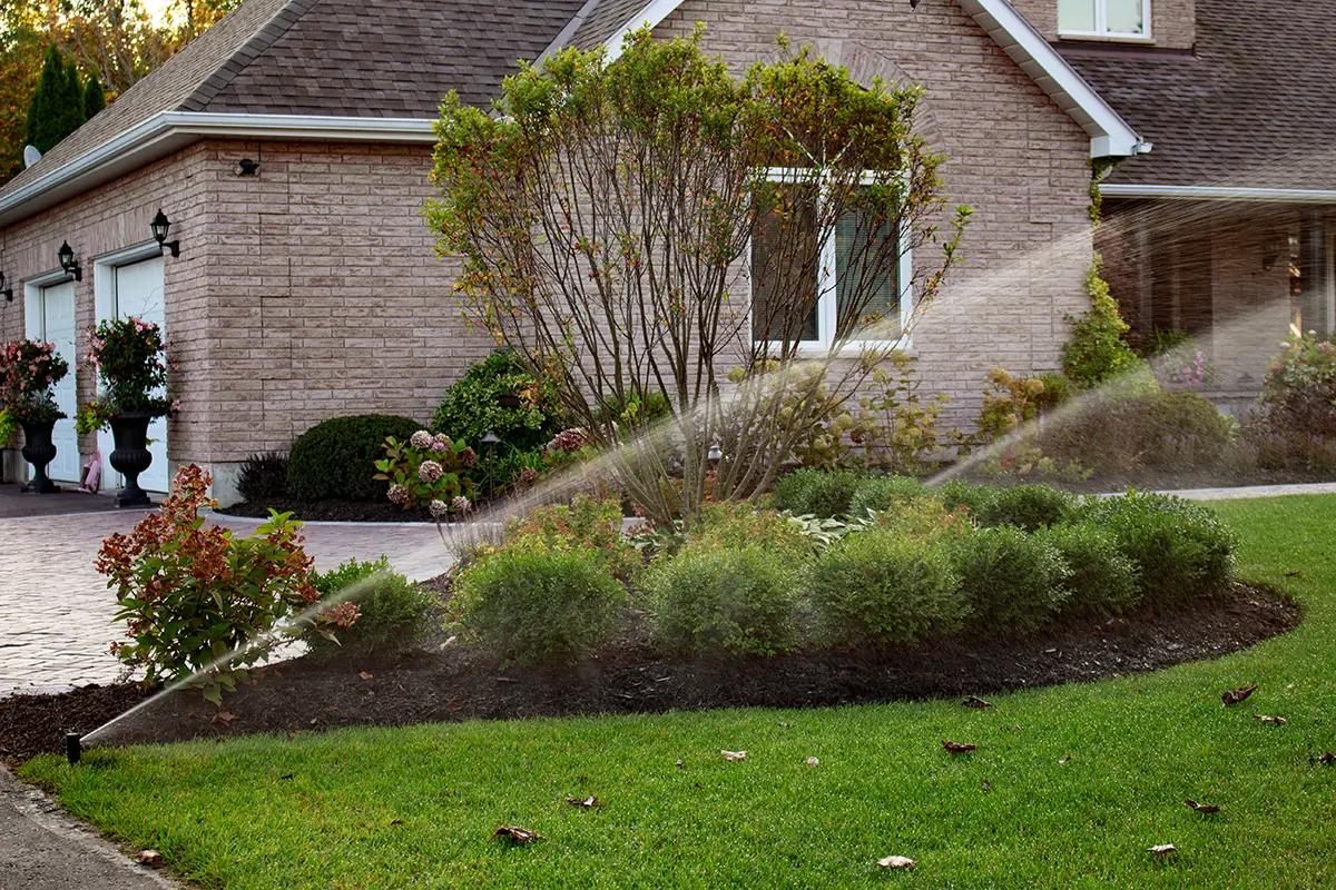 Two spray heads watering a mulched foundation bed and lawn in front of a brick house.