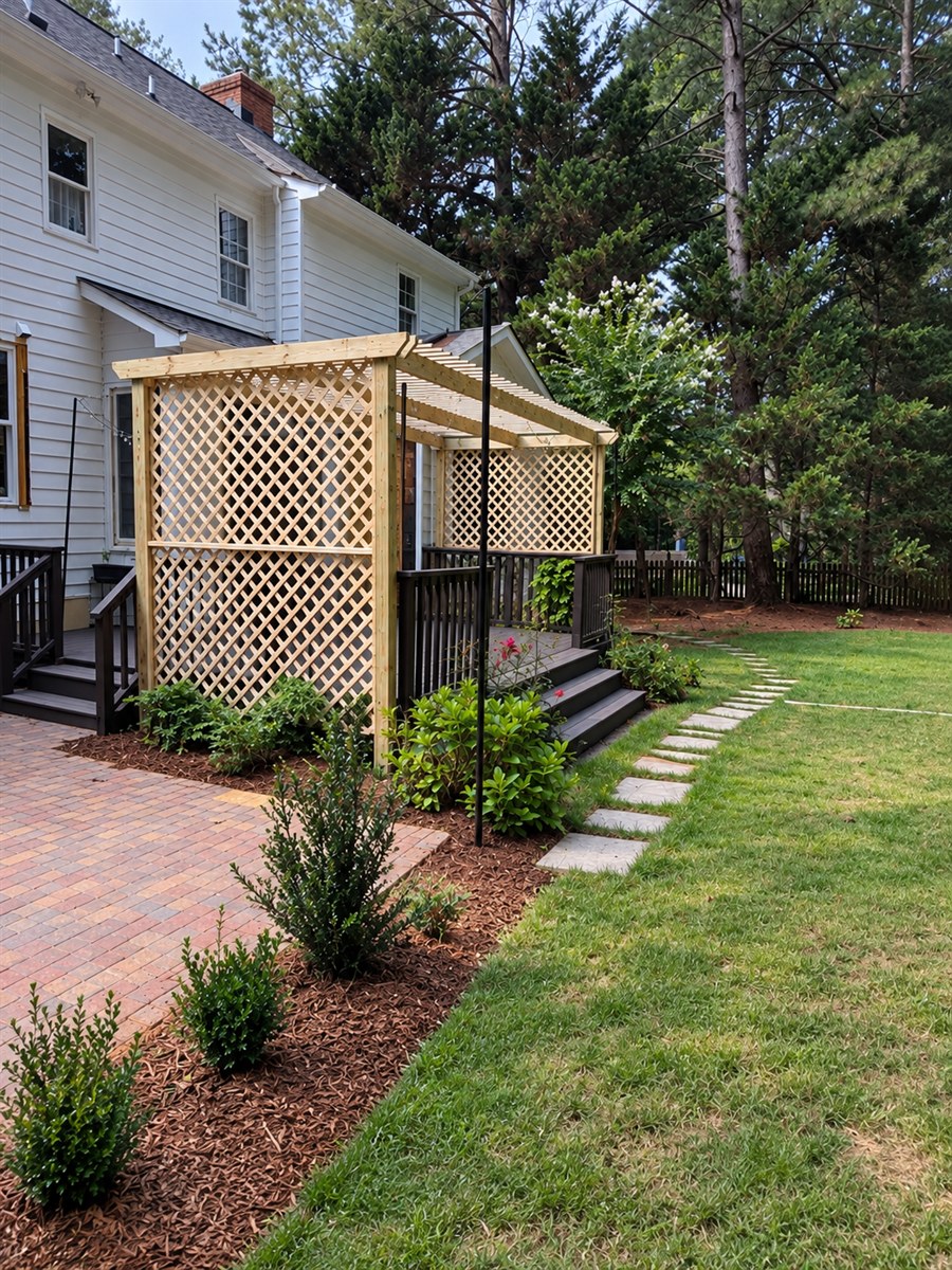 A wooden lattice vine trellis with an open overhead beside a dark-stained deck, young shrubs in fresh mulch along a brick patio, and stepping stones across the lawn.