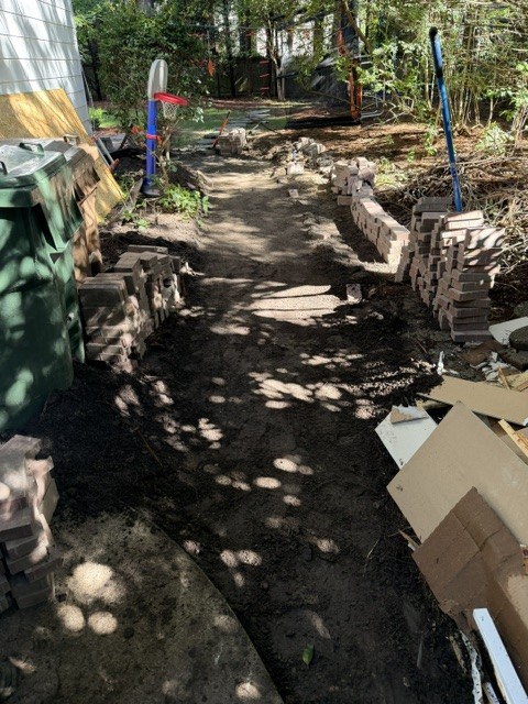 A narrow side yard excavated to subgrade, with brick pavers stacked in rows along both sides ready to lay.